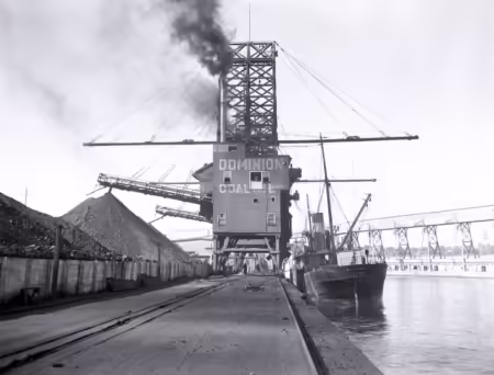 A black-and-white photo of an industrial structure bearing the words "Dominion Coal" on a long pier. The building has booms extended over piles of crushed coal to the left and a ship on the right. Black smoke rises from the building.