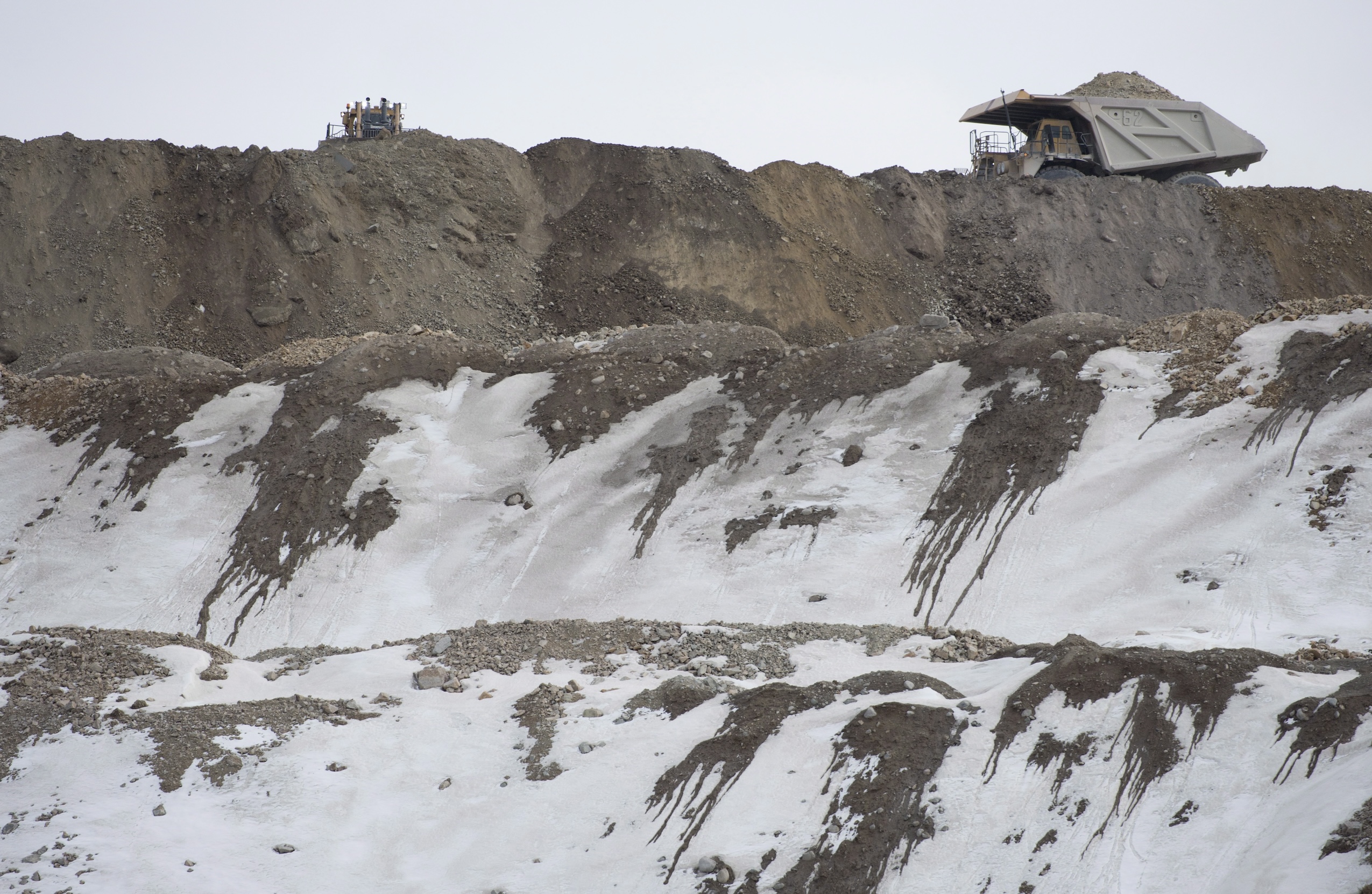A large dump truck is perched at the top of several tiers of a mine excavation, the bare ground partially covered in snow