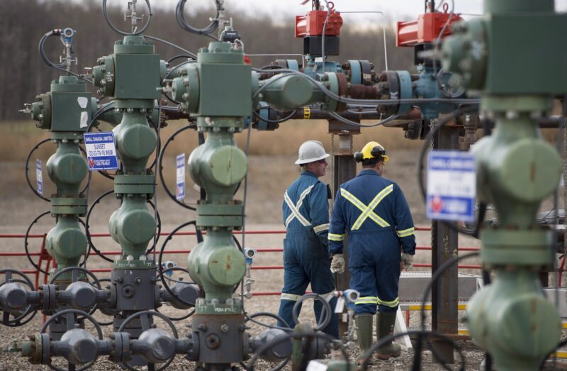 two workers in hard hats and coveralls walk near industrial pipes and natural gas infrastructure