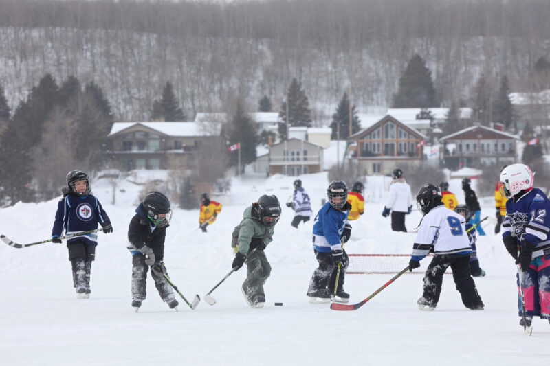 Young children in hockey jerseys and warm winter gear play hockey on the ice on a wintry day