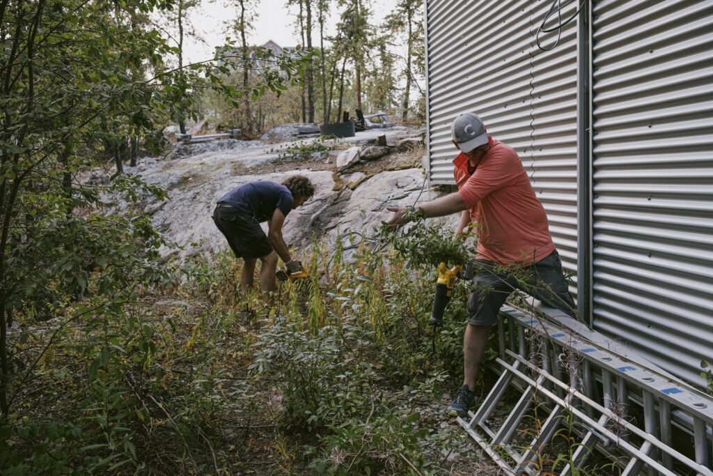 Two men use small power tools to clear vegetation growing against the siding of a building