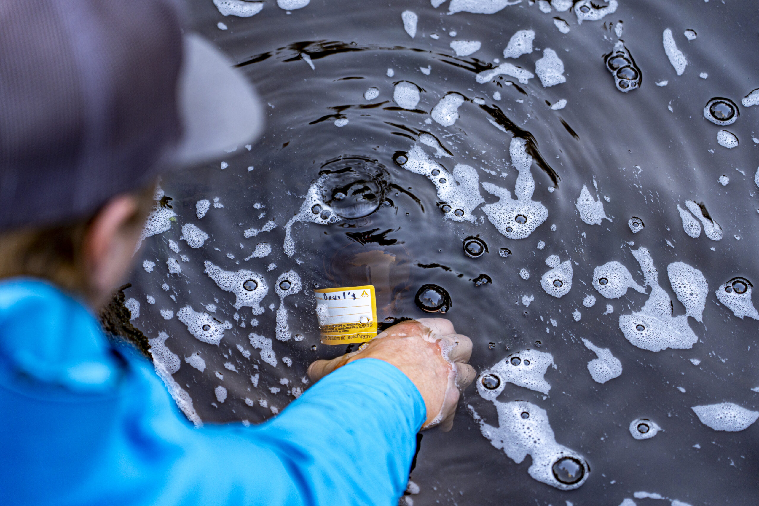 A man in a blue jacket holds a sampling bottle labelled 'Devil's' under water with foam at the surface