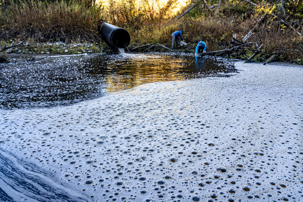 Two men bend down taking samples at the shore of Blackbird Creek, next to a black pipe pouring into the water where foam and bubbles coat the surface