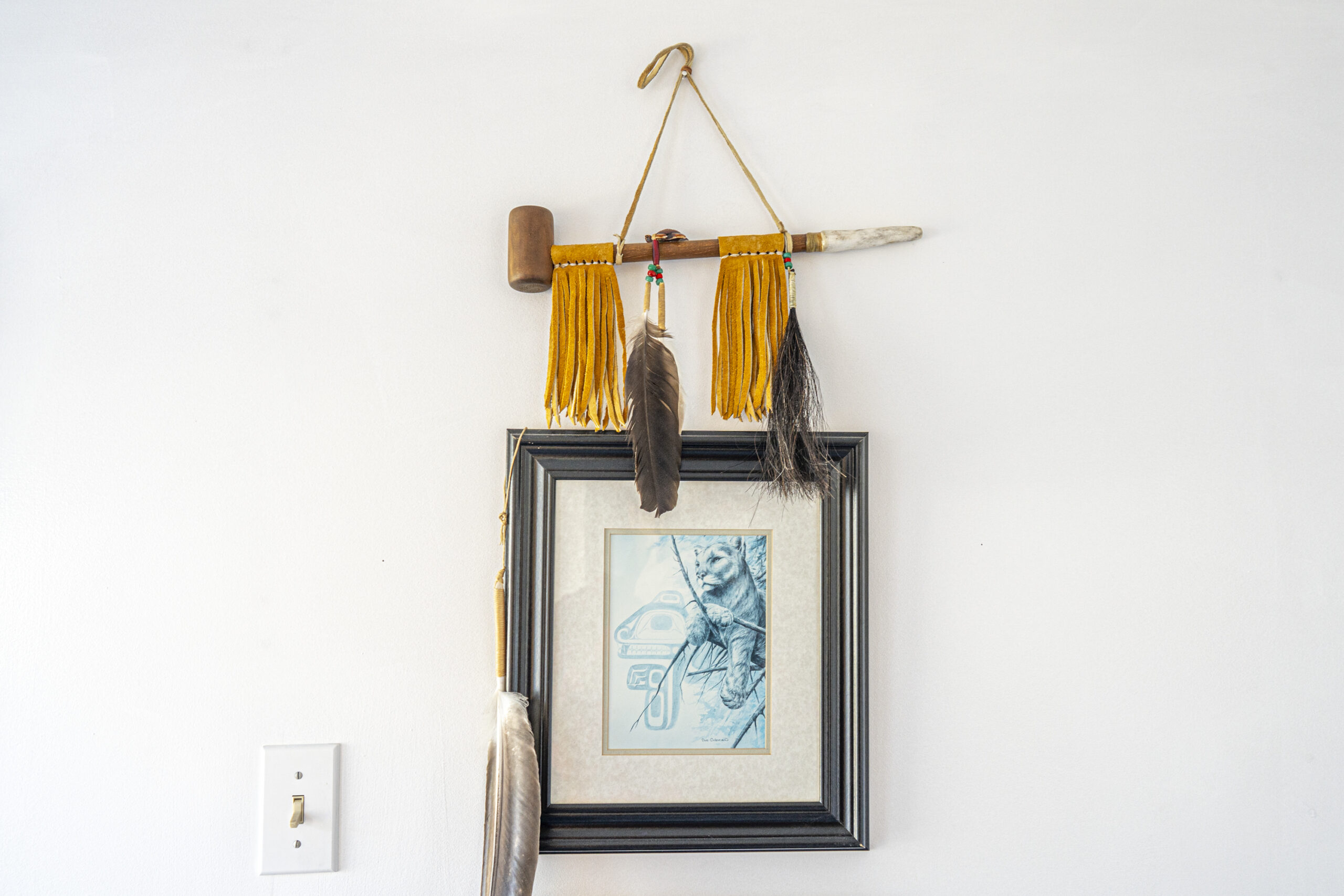 A picture of a mountain lion hangs on a white wall with hide tassels and feathers hanging from a ceremonial pipe above it