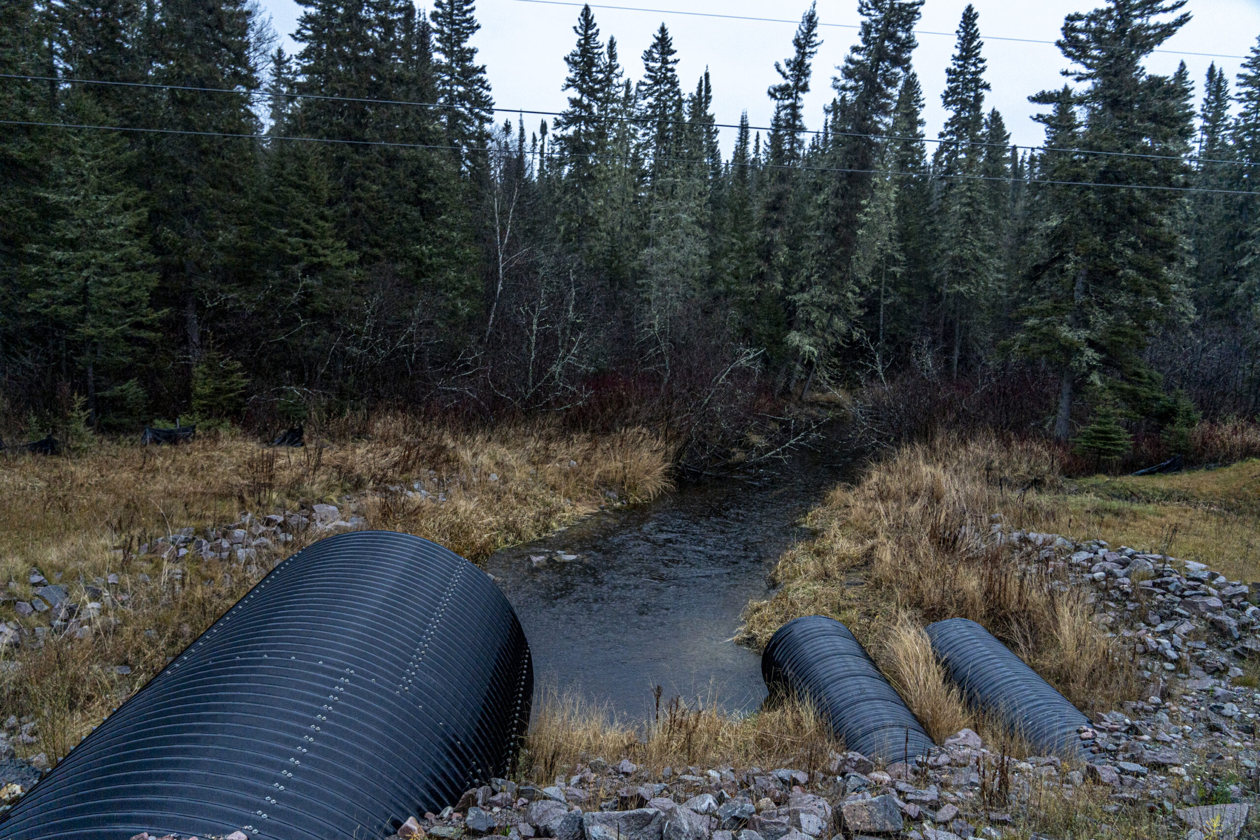 Three black pipes stick out of the ground, aimed at a creek with trees behind it