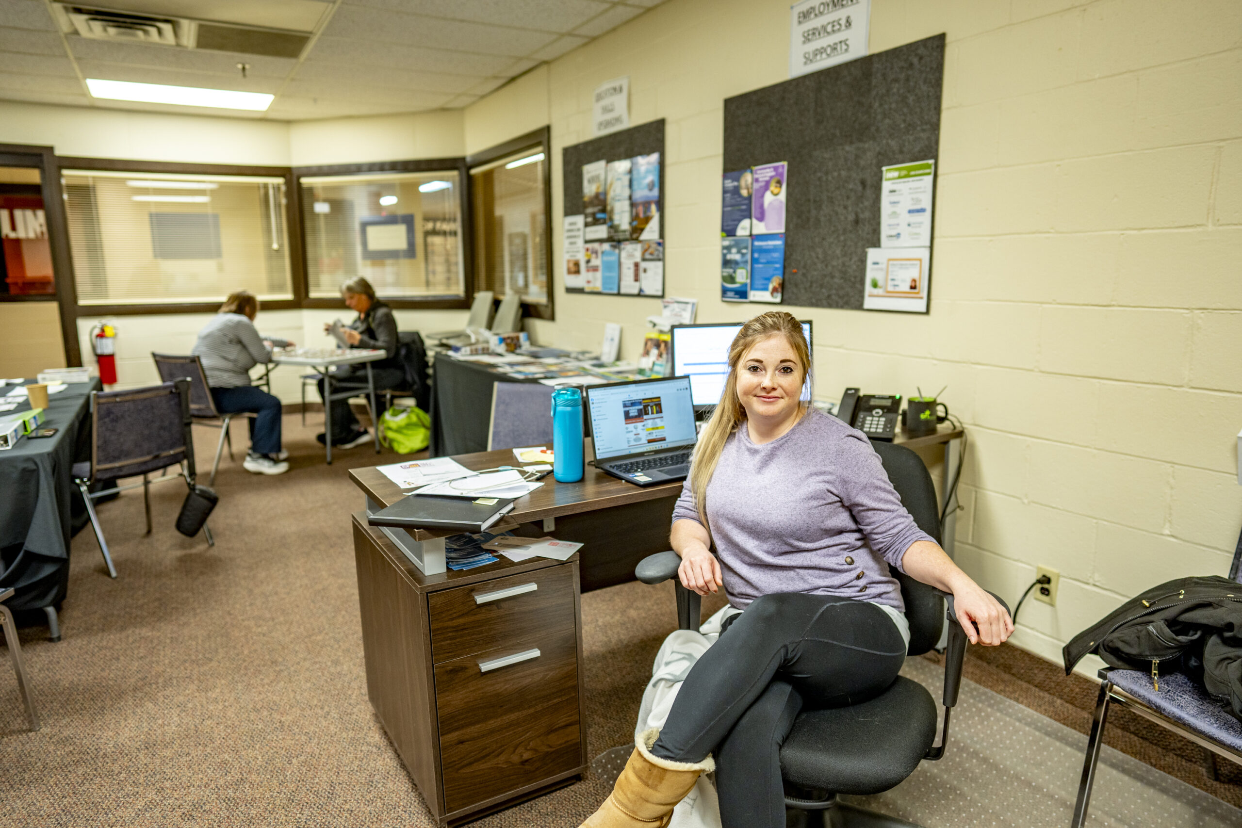 A woman sits at a chair in front of a desk in an office where other people sit at a table behind her