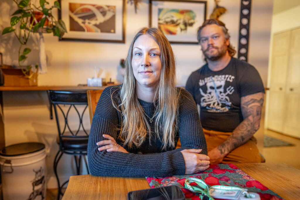 a woman sits at a table looking toward the camera with a man sitting off to her left, also looking at the camera