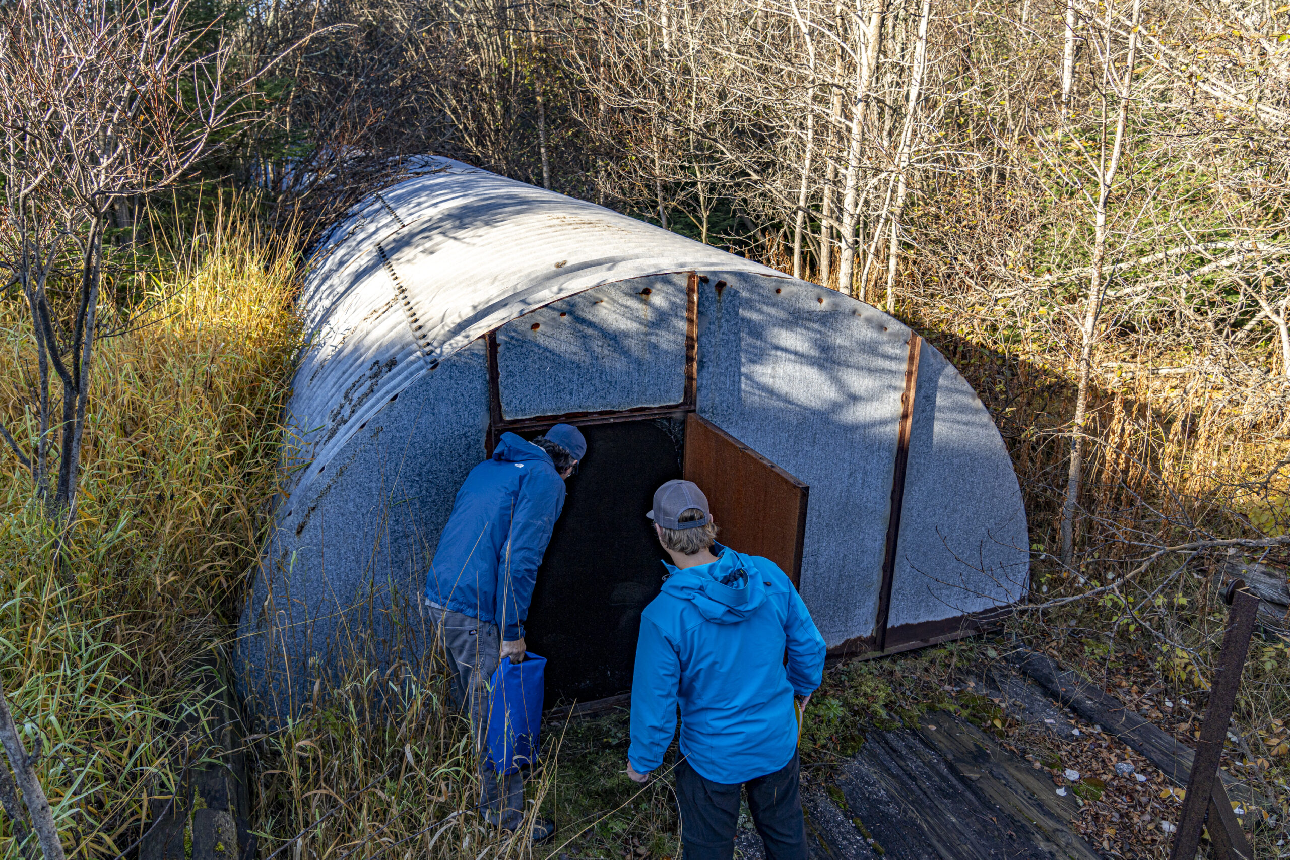 Two men look into the open door of a low quonset hut surrounded by brush