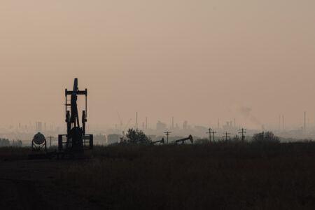 A hazy, dark sky and darkened field in Alberta with a pump jack in the foreground and an industrial area in the background. The industry is overseen by the Alberta Energy Regulator.