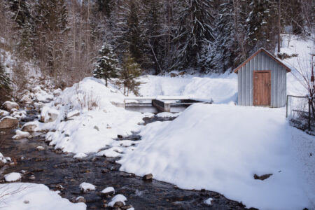 A small creek flows near snow-covered banks, trees and a small shed