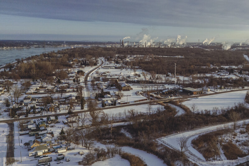 Smoke billows from refineries in the distance with trees, roads and houses in front of it