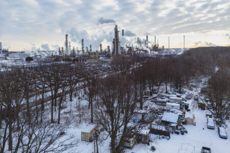 Smoke pours out of refinery towers beyond a cluster of sheds and a few rows of trees