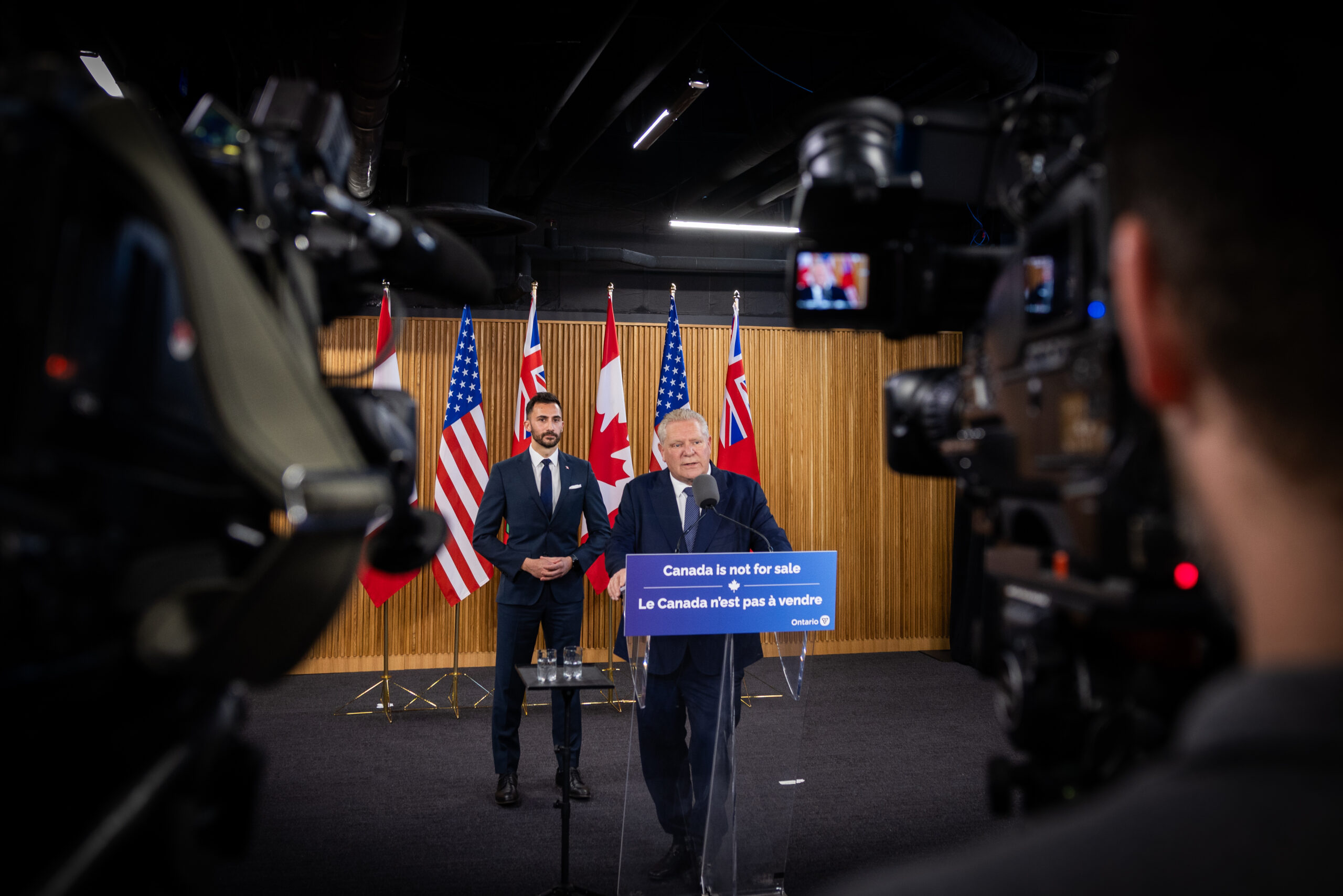 Ontario Premier Doug Ford stands at a lectern reading 'Ontario is not for sale' with Energy Minister Stephen Lecce standing behind him and various cameras pointing at them