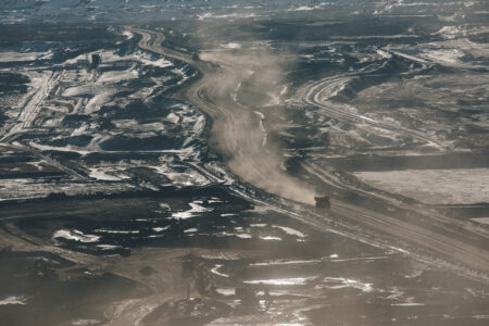 An aerial shot of an oilsands mine, with a giant mining truck, driving down a roadway looking very small.