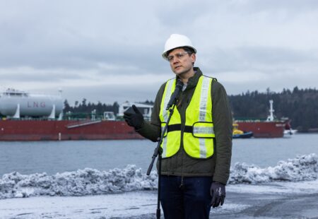 David Eby, wearing a hard hat, reflective vest and safety glasses, holds up a hand while speaking at a microphone. A large ship behind him has a tank with "LNG" painted on it.