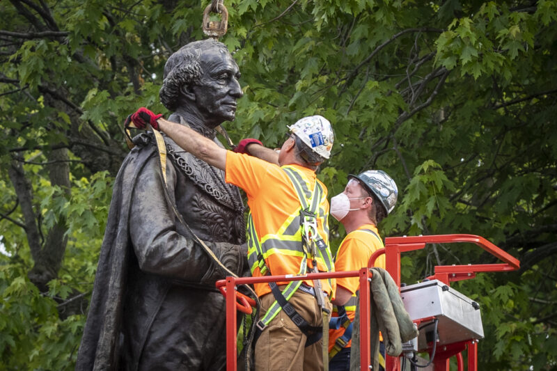 Workers on a cherry picker in high-vis wrap straps around a statue of John A. MacDonald