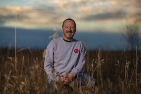Drew Anderson wears a Narwhal-branded sweated and kneels among Prairie grass, smiling