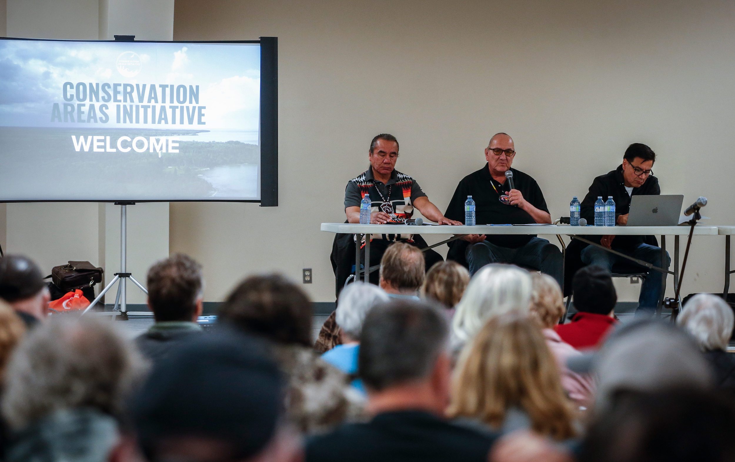 Chief Tony Travers, Kinonjeoshtegon First Nation, from left, Chief David Crate, Fisher River Cree Nation, and Chief Stan Bird, Peguis First Nation sit on a panel next to a screen reading "Conservation Areas Initiative Welcome"
