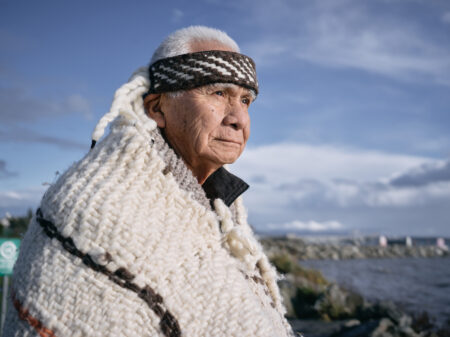 WSANEC Chief Vern Jacks looks off camera to the right, sun glowing on his face, wearing white and brown wool regalia, with a blue sky behind him