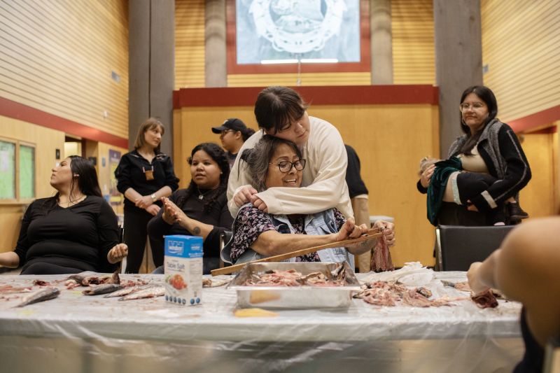 Elder Roseanne George shares a moment with Brooklyn Baker as she prepares herring fish in Totem Hall, with other members of the nation behind her