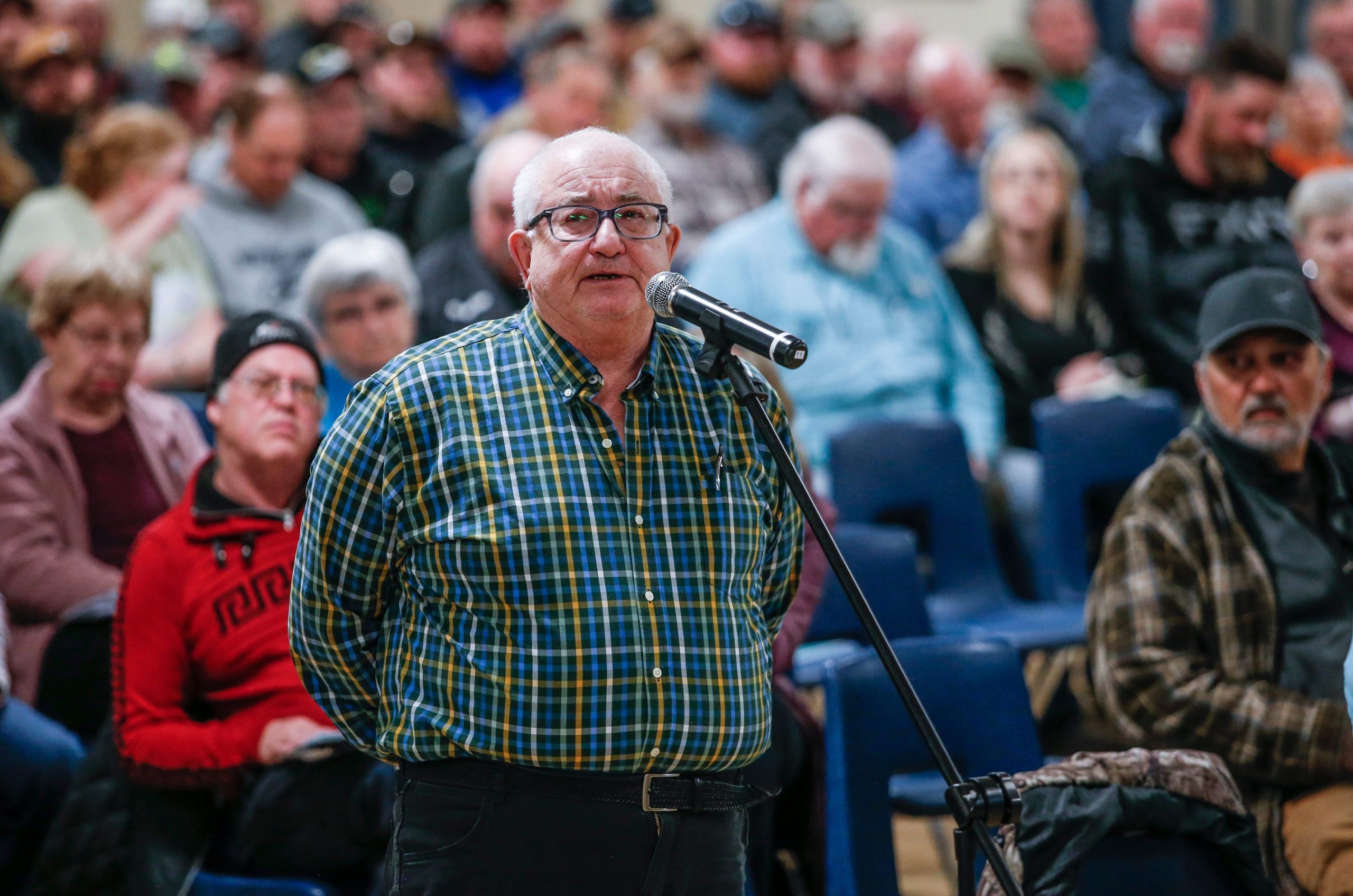 Dennis Schindler wears a blue and yellow checked shirt as he stands at a microphone in front of a crowd of people in Arborg, Manitoba to speak about a conservation area proposal