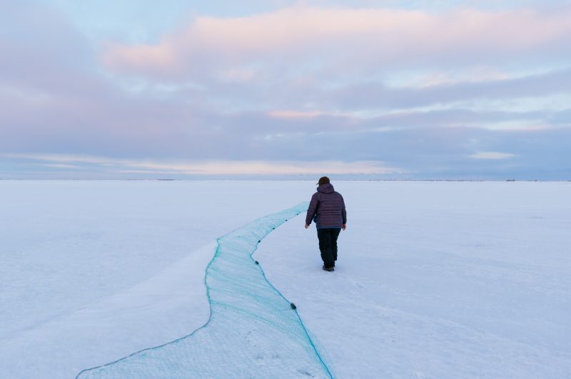 A man walks along a fishing net laid across the ice under a cloudy sky