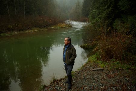 Katzie councillor Rick Bailey stands on the bank of the Alouette River, looking off camera to the left, wearing a green coat. Behind him, the river water is low, slow and green. Mist hangs over the trees extending behind him.