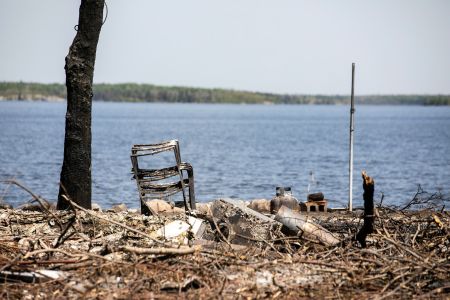 The aftermath of a fire, including a stack of burned camp chairs, a charred tree trunk and debris, near a lakeshore.