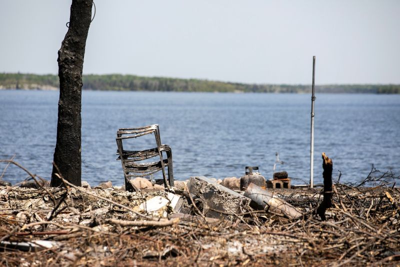 The aftermath of a fire, including a stack of burned camp chairs, a charred tree trunk and debris, near a lakeshore.