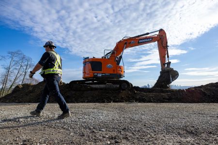 A man in a high-vis vest and hard hat walks past an excavator that is shovelling gravel