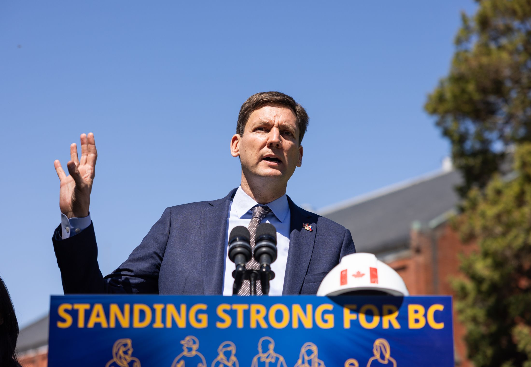 B.C. Premier David Eby stands behind a lectern with a sign that reads, "Standing Strong for BC" in yellow text on a blue background. He is wearing a blue suit jacker, shirt and tie. There is a white hard hat with a Canada flag on the lectern to his left