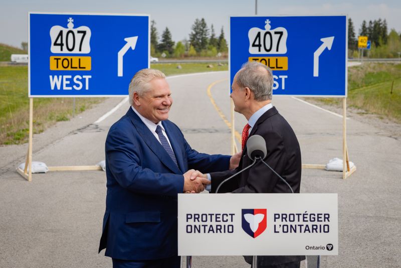 Ontario Premier Doug Ford and Finance Minister Peter Bethlenfalvy shake hands at a podium that reads 'Protect Ontario,' standing on a road in front of signs for Highway 407