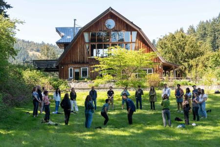 Two dozen people stand in a circle in a grassy area, apparently playing a game, in front of a large, wooden, barn-shaped building