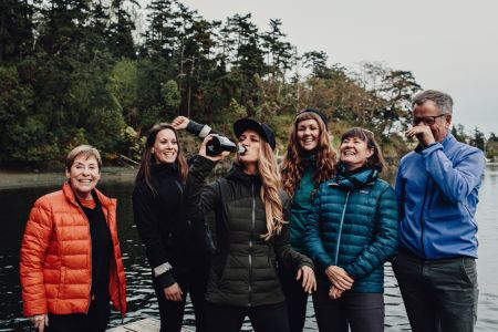 Six people smile and laugh on a dock. One is drinking from a champagne bottle, one raises an arm in a fist pump.