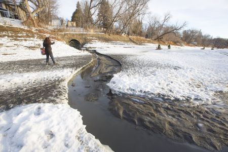 A woman stands near a storm drain, with visible brown runoff flooding into a frozen river