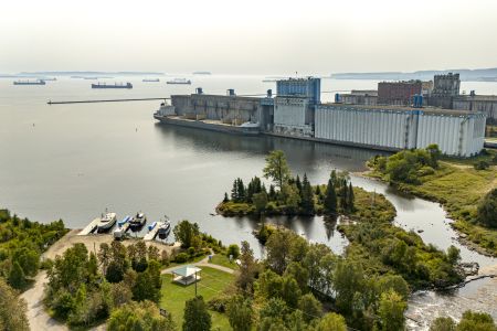 An aerial view of an industrial ship docked by a grain elevator by a park. Other ships can be seen in the horizon, with an island further in the distance in the mist.