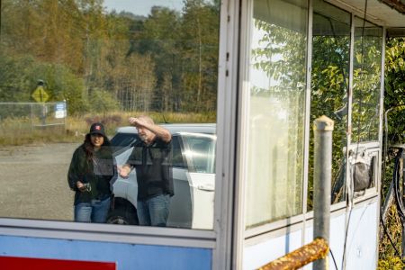 The reflection of two people standing next to a car is seen through glass windows of a small structure at the entrance to an industrial site.