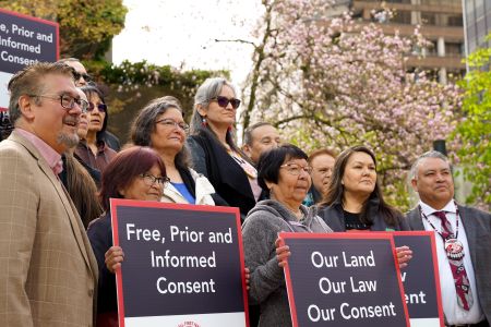 A group of Xatśūll community members and allies stand as a group, facing to the right, looking off camera, holding signs that say "free, prior and informed consent" and "our land, our law, our consent"