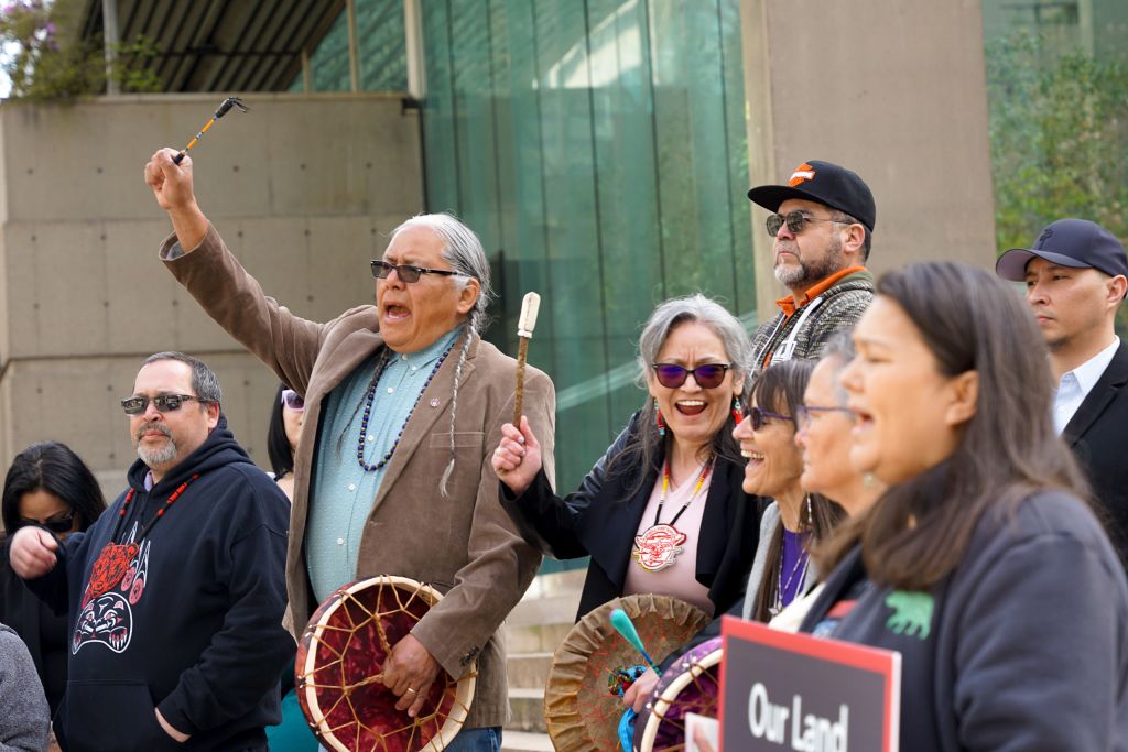 A group of people sing, some holding drums or signs, all facing to the left. In the centre, Chief Rhonda Phillips smiles with the person next to her, mouth open singing, and with her hand raised in the air, holding a drumstick.