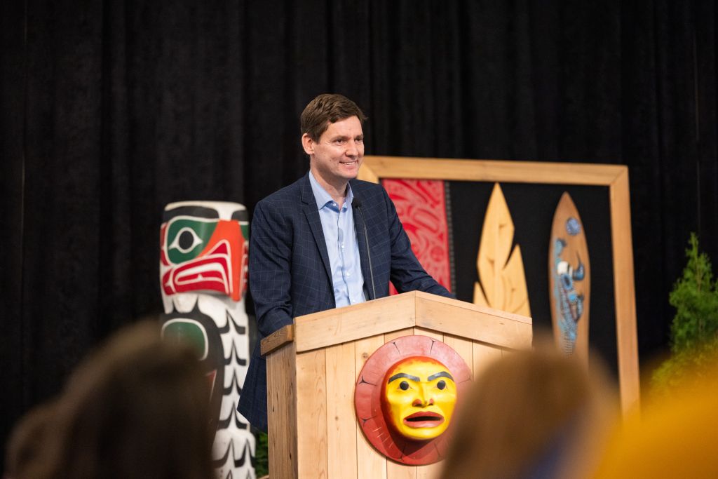 Premier David Eby stands at a pine lectern decorated with a First Nations mask. He's wearing a black suit and light blue shirt and tie. He's smiling, addressing a crowd