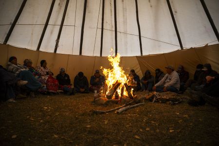 A dozen people sit around a large fire at the centre of a teepee