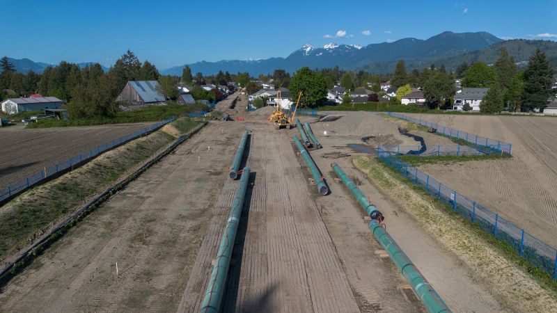 An aerial shot, looking down the length of a pipeline construction site, with pipes on the ground, cleared earth and mountains in the background.