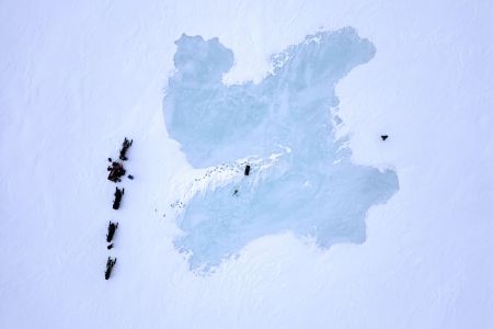 Aerial view over snow-covered sea ice. A patch in the middle is darker and snow-free. People and snowmobiles gather to the side.