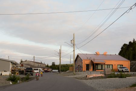 In Bella Bella the sky is soft and pale blue, with warm light illuminating houses and a larger building covered in Heiltsuk art. Two children ride their bikes down the wide open street