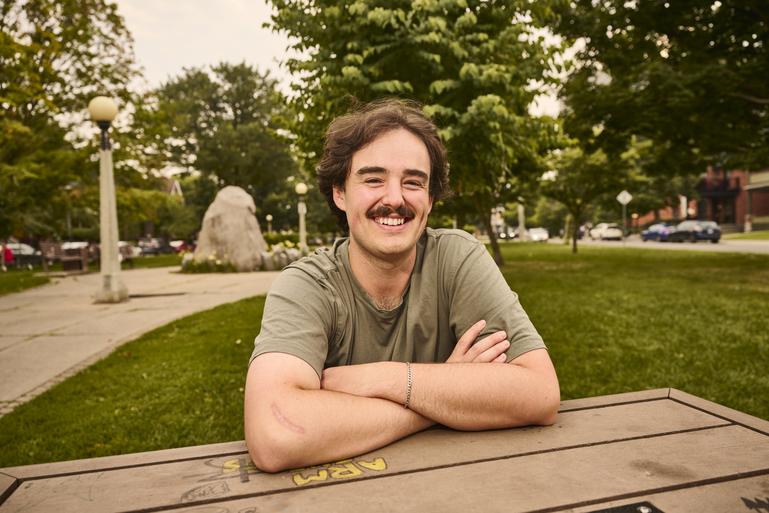 A man with a moustache sits at a picnic table with his arms crossed, smiling into the camera.