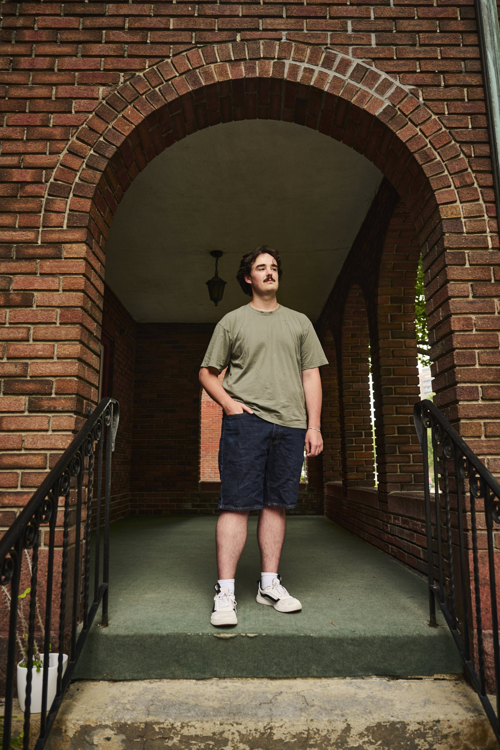 A man in a t-shirt stands under a brick archway, with one hand in a pocket.