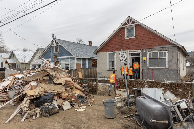People in high visibility vests enter a small home. There is debris piled in the yard outside