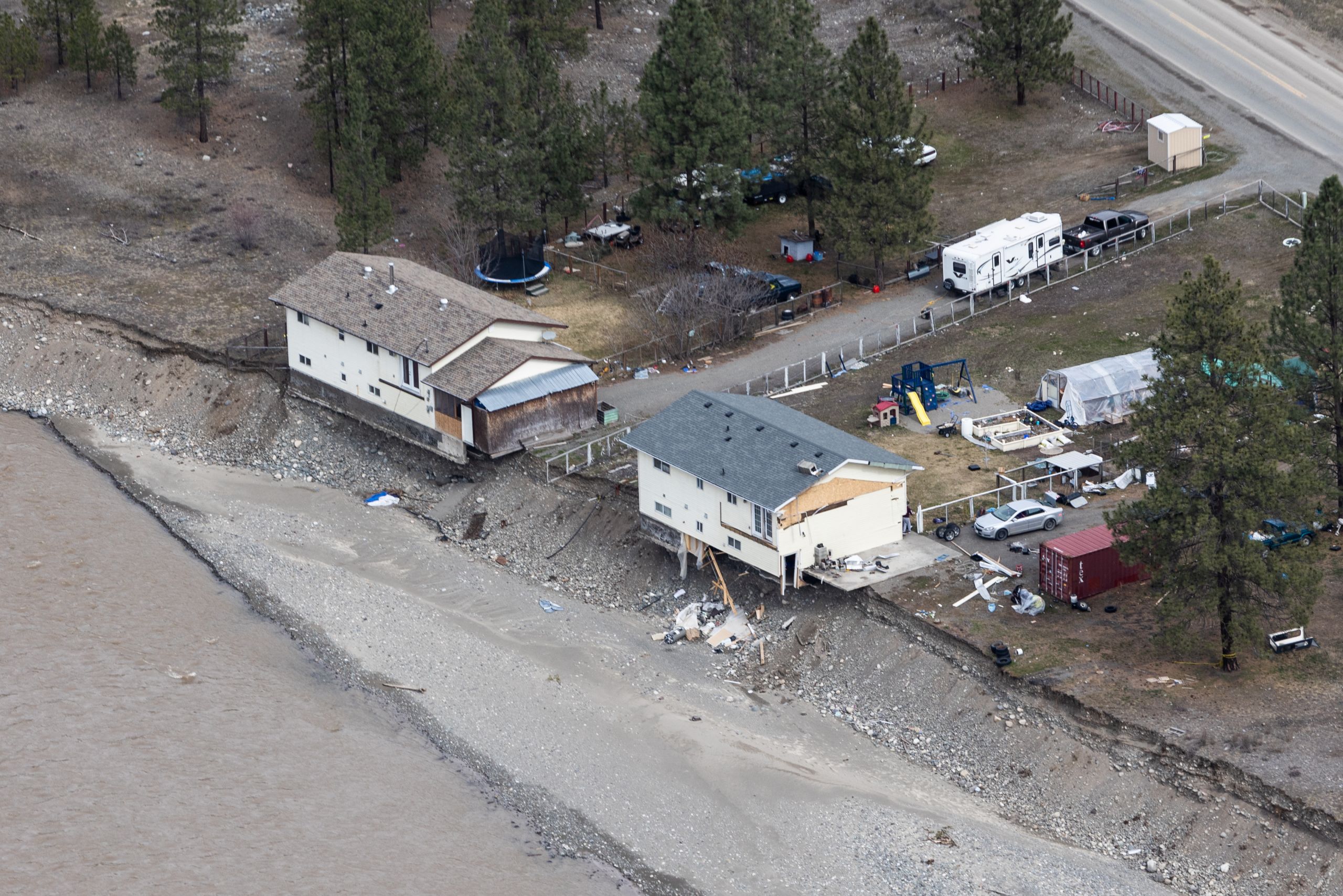 Two homes sit precariously on a bank that has clearly been carved out by water. There is debris in front of the homes and behind them, below the bank