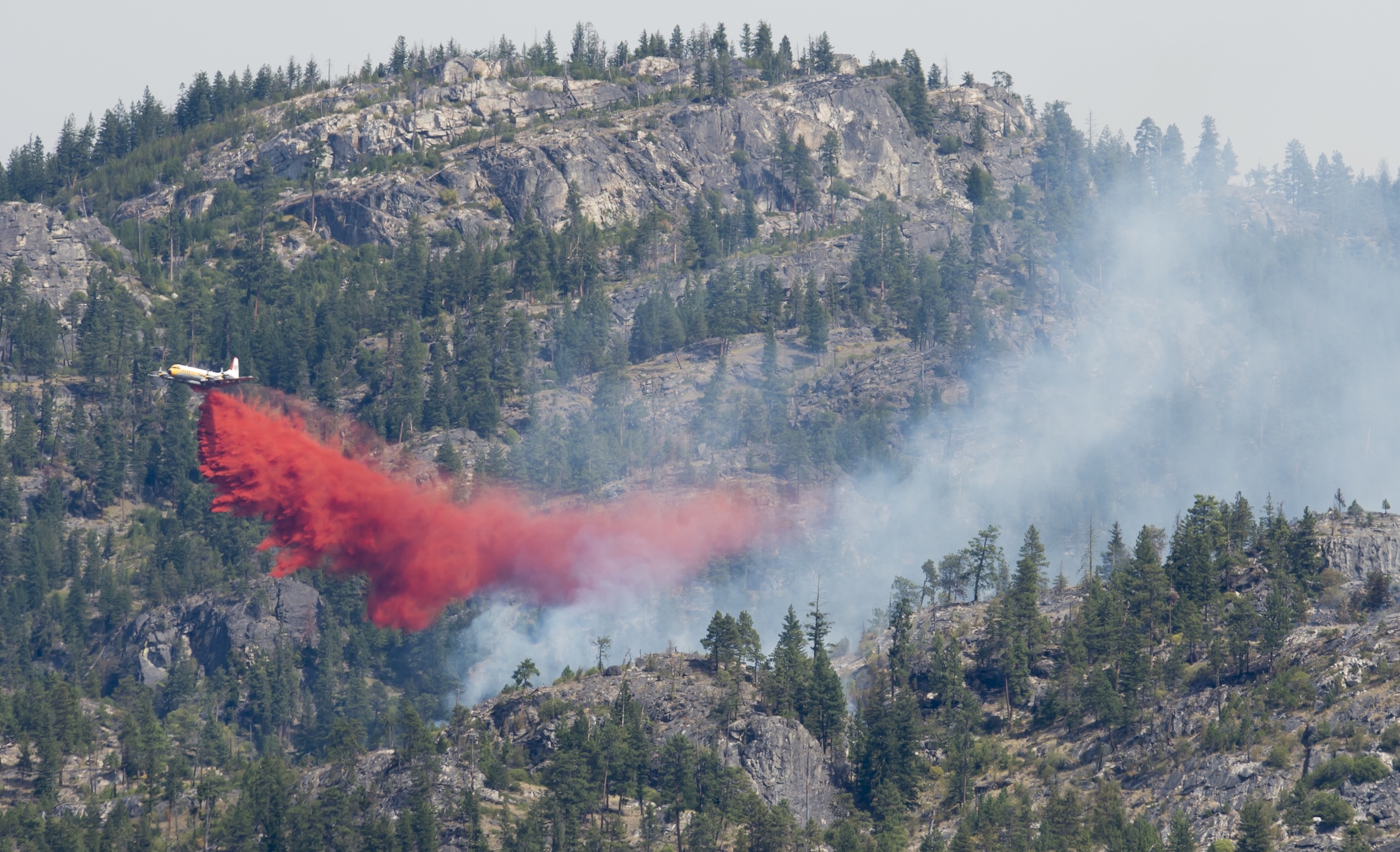 A plane releases a bright red plume of fire retardant as it flies over a rocky, mountainous area.