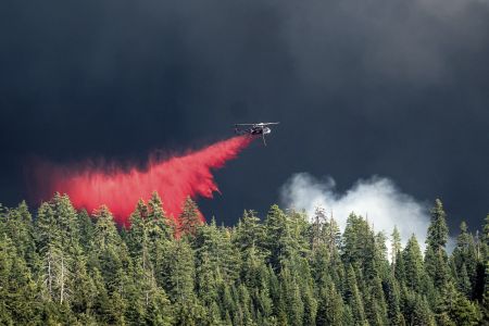 A helicopter flies over a stand of trees, dropping a bright red plume of fire retardant.